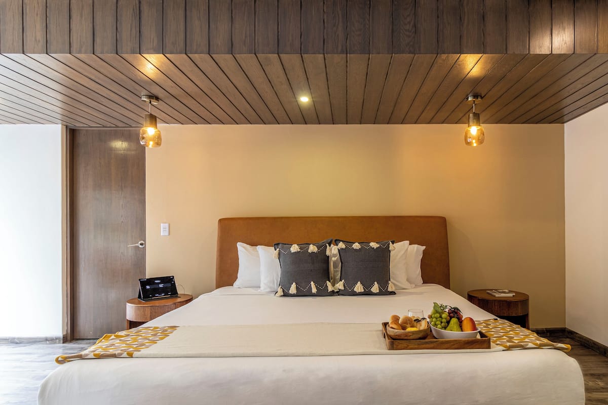 A hotel room with a wooden ceiling, a large bed with a headboard, bedside tables, and a tray with fruits and bread.