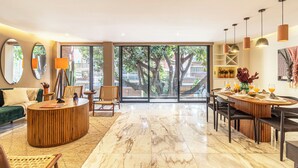 A spacious living area with a round wooden coffee table, a green sofa, and a dining area with a wooden table and chairs. Large windows provide a view of trees and buildings outside. - Hotel Boutique Kooben  (Merida)