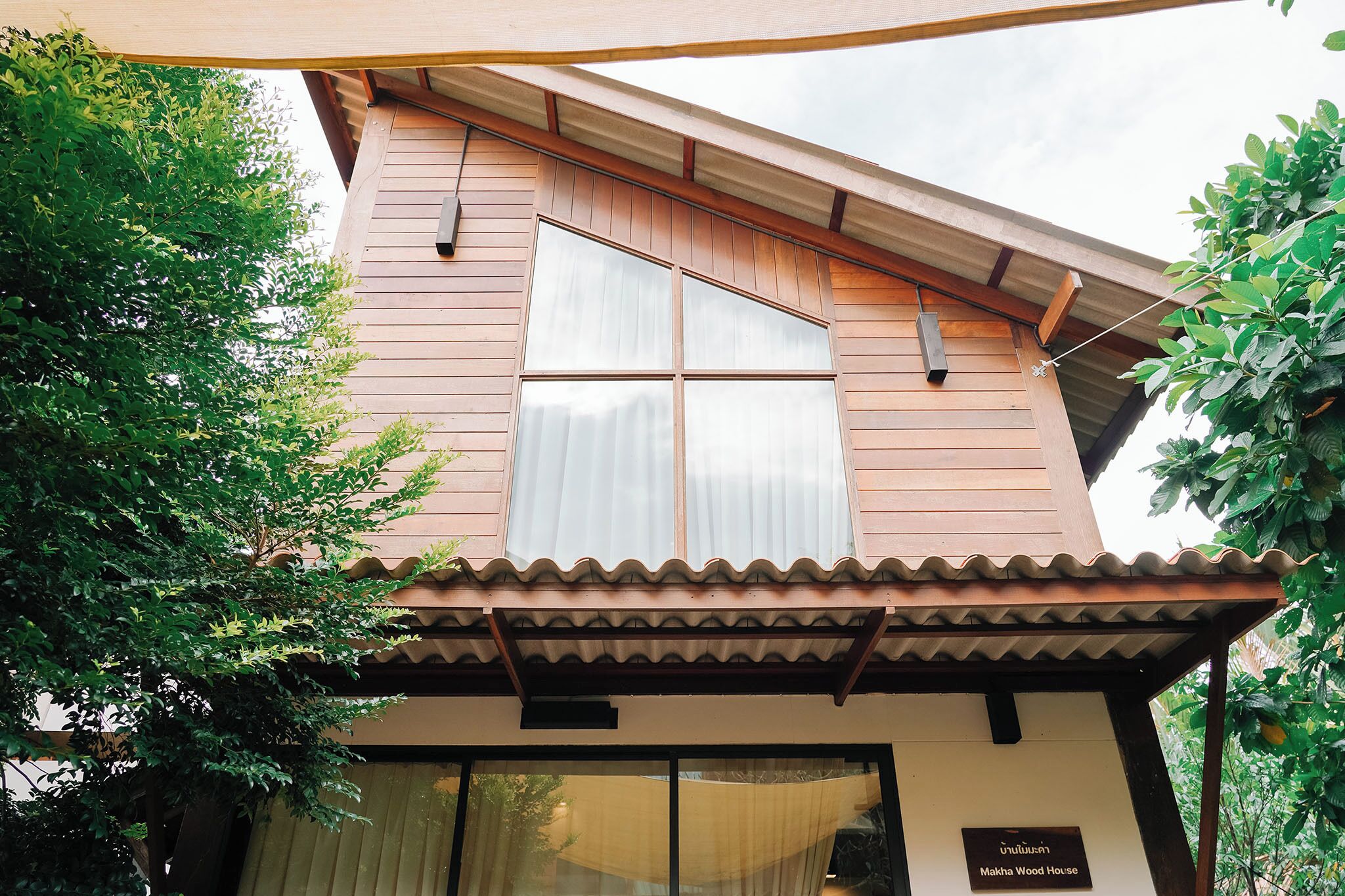 A two-story wooden house with a large window and a sign reading "Makha Wood House".