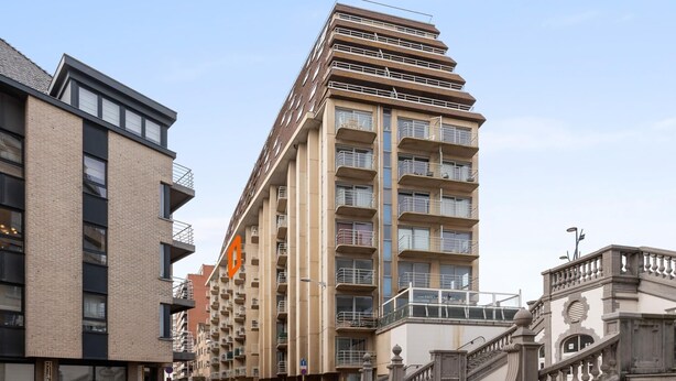 Un edificio moderno de varios pisos con un logotipo naranja llamativo, una escalera con barandillas ornamentales y una plaza empedrada con ladrillos.