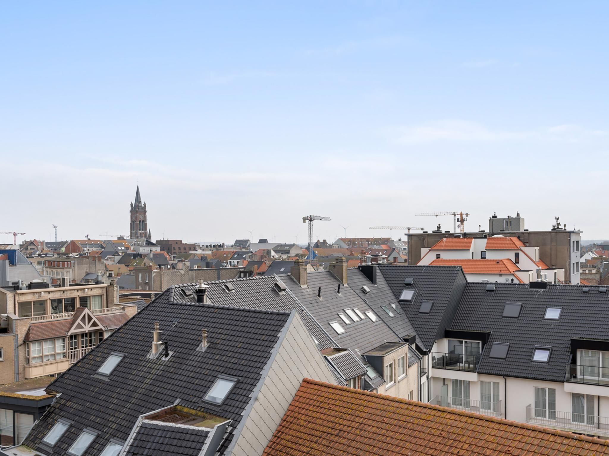 Une vue urbaine avec de nombreux toits, dont certains sont dotés de lucarnes et de fenêtres en lucarne, et un clocher d’église imposant en arrière-plan.