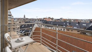 Un balcón con mesa y sillas, con vistas a un paisaje urbano que incluye varios edificios y la aguja de una iglesia a lo lejos.