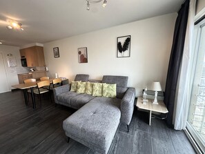 A modern living room with a grey sofa, a dining table, and a kitchen in the background. - Sunny apartment with sea-views (Blankenberge)