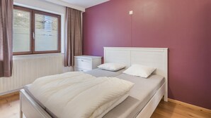 A bedroom with a bed, a nightstand, a window with curtains, and a wall painted in a solid color. - Pleasant apartment near the beach (Blankenberge)