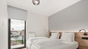 A modern bedroom with a large bed, a wooden headboard, a bedside table, and a view of the city through a large window. - Beautiful apartment in the center (Blankenberge)