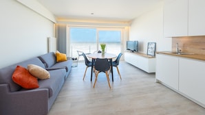 Ein modernes Wohnzimmer mit grauem Sofa, einem Esstisch mit Stühlen, einem Fernseher und einer Küchenzeile mit weißen Schränken und einer Holzarbeitsplatte.