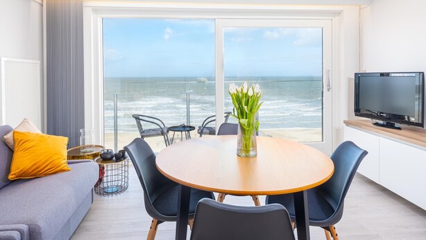 Ein modernes Wohnzimmer mit großem Fenster mit Meerblick, einem runden Holztisch mit Stühlen, einer Couch mit Kissen und einem Flachbildfernseher auf einem Holzboden.