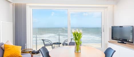 Una sala de estar moderna con un gran ventanal con vistas al océano, una mesa redonda de madera con sillas, un sofá con cojines y un televisor de pantalla plana sobre un mueble de madera.