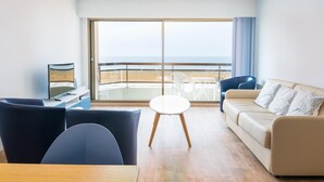 A modern living room with a sofa, chairs, a small table, and a TV. It features a large sliding glass door leading to a balcony with a view of the beach. - Modern apartment with direct sea view (Middelkerke)