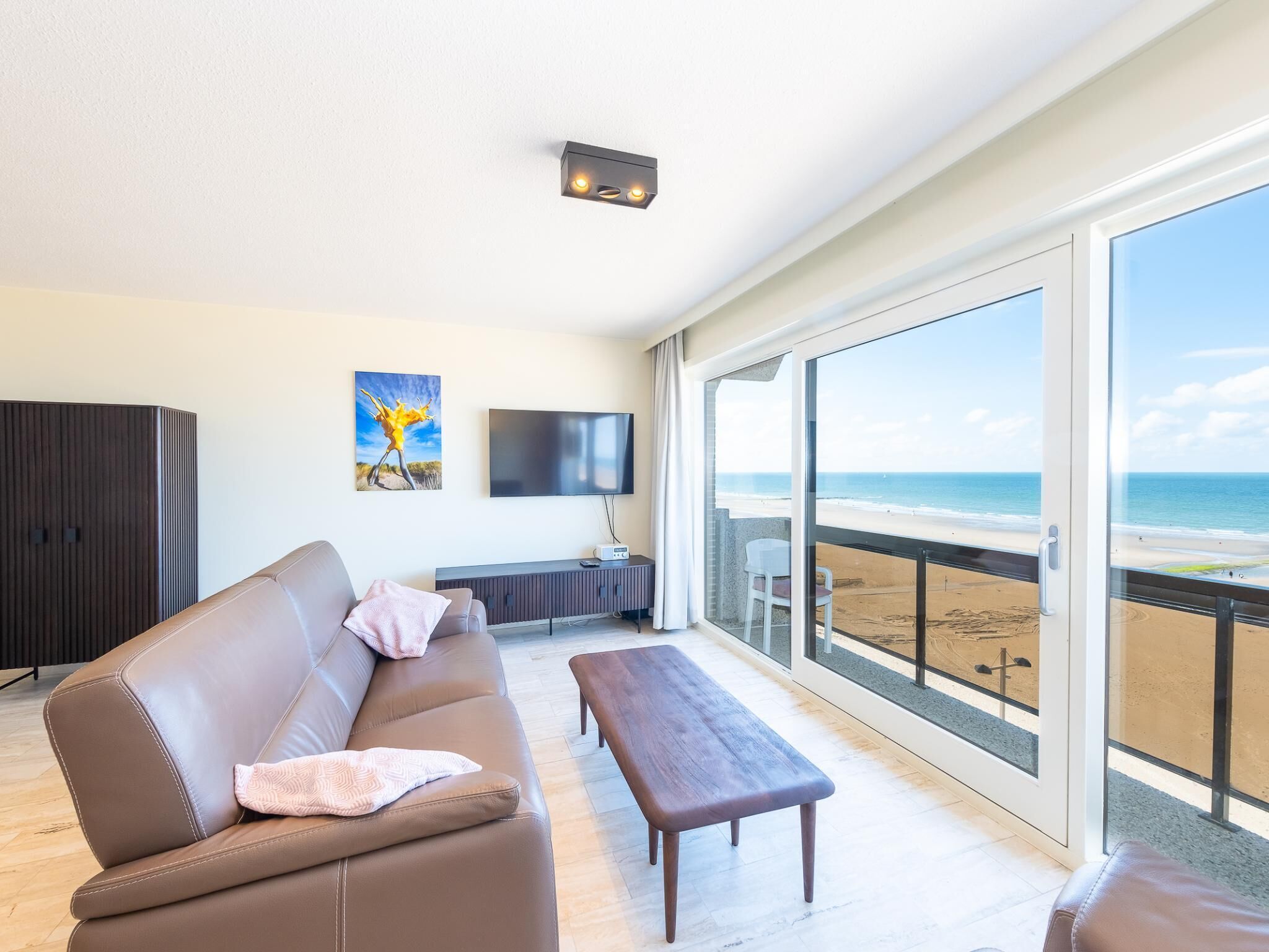 Una sala de estar moderna con un sofá, un banco, un televisor y un amplio ventanal con vista a una playa.