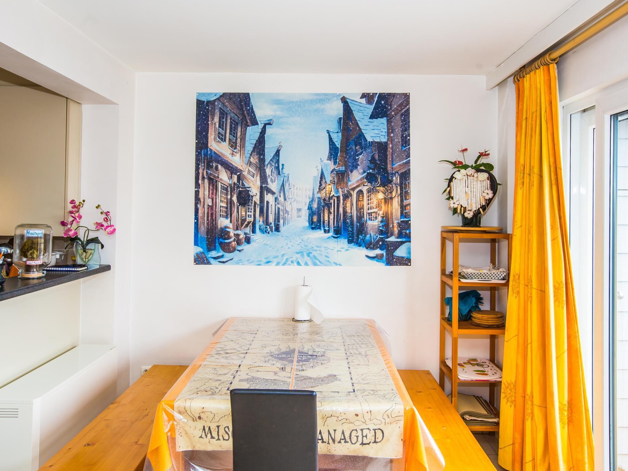 A dining area with a table covered by a map-themed tablecloth, a painting of a snowy street on the wall, and a wooden shelf with decorative items.