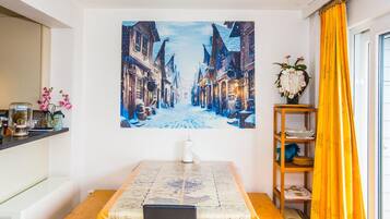 A dining area with a table covered by a map-themed tablecloth, a painting of a snowy street on the wall, and a wooden shelf with decorative items.