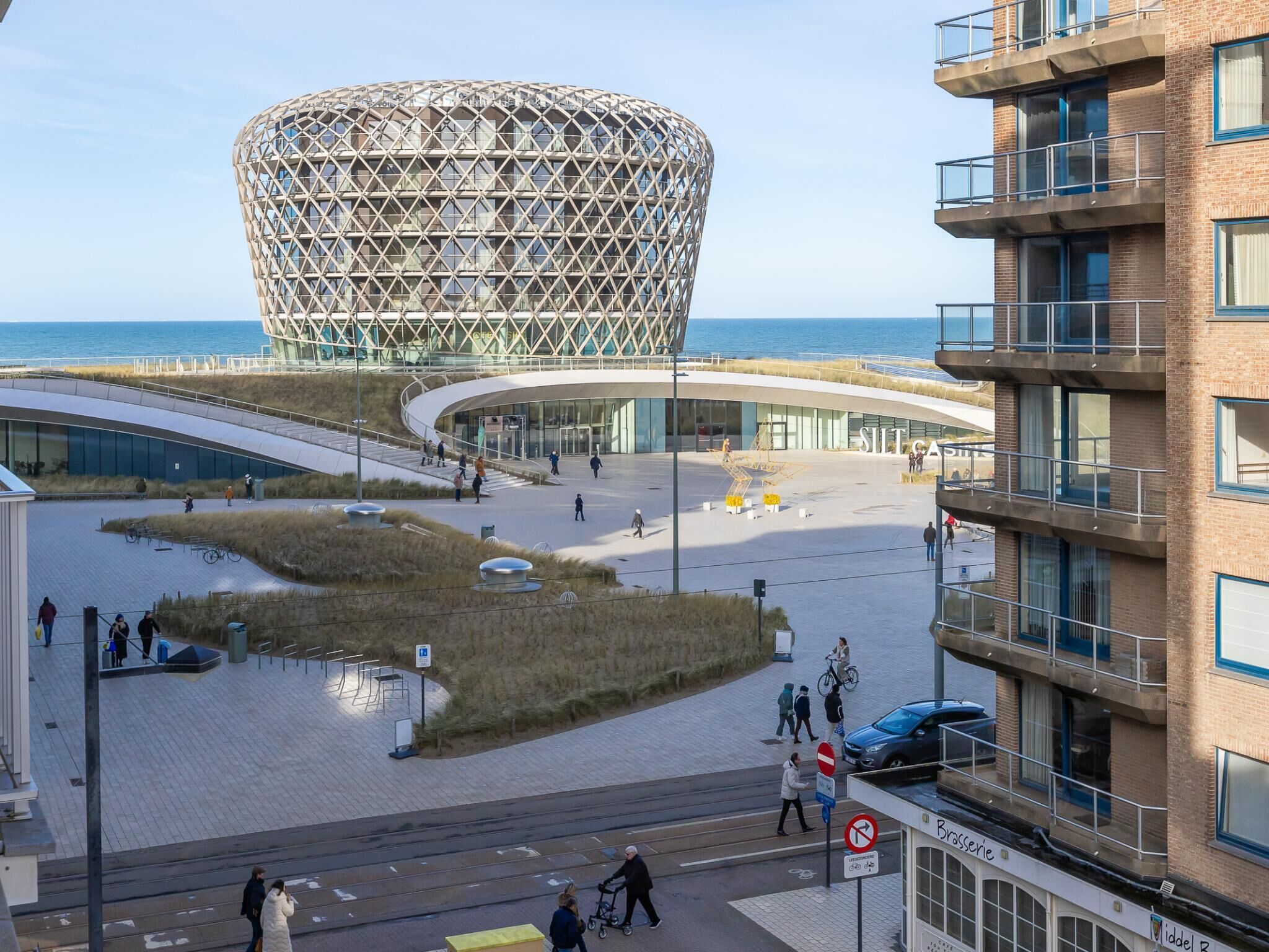 A modern building with a unique geometric facade, a pedestrian area with benches, and a view of the sea.