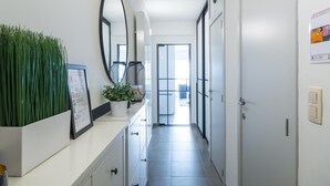 Ein Flur mit einer weißen Theke, einem Blumentopf, einem großen Spiegel und einer Tür zu einem hellen Raum.
