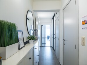 Ein Flur mit einer weißen Theke, einem Blumentopf, einem großen Spiegel und einer Tür zu einem hellen Raum.