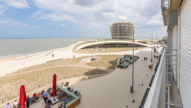 Eine Uferpromenade mit einem markanten, modernen Gebäude, einem Sandstrand und Außenbereichen zum Verweilen.