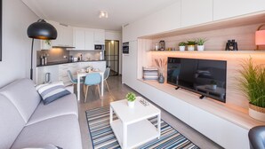 Ein modernes Wohnzimmer mit Sofa, Couchtisch und Fernseher. Es gibt eine Essecke mit Stühlen und einem Tisch. Im Hintergrund ist eine Küche zu sehen.