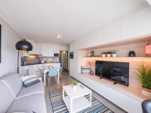 Ein modernes Wohnzimmer mit Sofa, Couchtisch und Fernseher. Es gibt eine Essecke mit Stühlen und einem Tisch. Im Hintergrund ist eine Küche zu sehen.