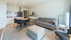 En moderne stue med en grå sofa, et rundt spisebord med stole, en lille skammel og en hvid radiator.