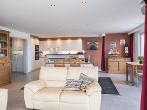 A modern living room with a beige sofa, a dining area with a wooden table, and a kitchen with white cabinets and recessed lighting. - Apartment with frontal sea-views (Middelkerke)