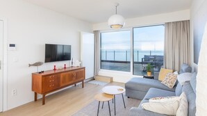 A modern living room with a large flat-screen TV, wooden cabinet, grey sofa, and a balcony with a view of the sea. - Exclusive penthouse with breathtaking 360° view (Middelkerke)