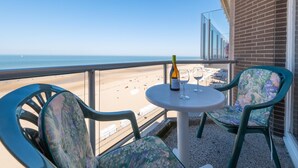 Un balcón con una mesa, dos sillas, una botella de vino y una copa. El balcón da a una playa y al océano.