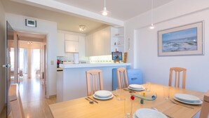 Un comedor moderno con una mesa de madera dispuesta para cuatro, una cocina al fondo y un cuadro en la pared.