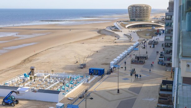 Un paseo marítimo frente a la playa con un edificio moderno característico, una playa de arena y un área de estacionamiento con una grúa azul.