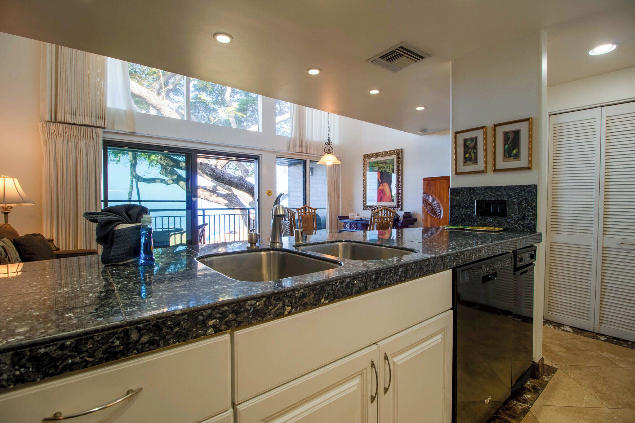 A kitchen with granite countertops, stainless steel sinks, and a dishwasher. There are two framed pictures on the wall, a window with curtains, and a ceiling fan.