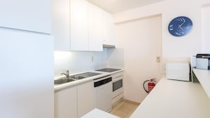 Cocina pequeña con gabinetes blancos, un lavamanos, una hornilla y un microondas. Hay un reloj en la pared y un extintor cerca de la hornilla.