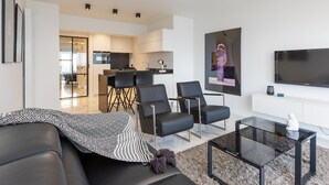 Ett modernt vardagsrum med en svart lädersoffa, ett soffbord i glas och en platt-TV. Längre bak i rummet finns en matplats med ett bord och stolar.