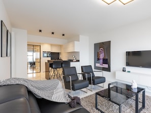 A modern living room with a black leather sofa, a glass coffee table, and a flat-screen TV. There is a dining area with a table and chairs in the background. - Luxury apartment with stunning sea views (Middelkerke)