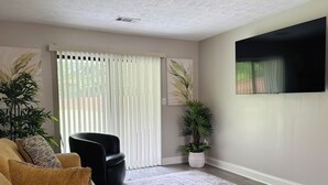 Ein Wohnzimmer mit einer Couch, einem schwarzen Sessel, einer großen Topfpflanze, einer Schiebetür mit Jalousien und einem Wand-Fernseher.