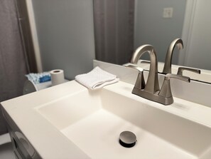 Un baño con un lavamanos blanco, dos llaves, una toalla y un rollo de papel higiénico.