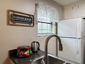 A kitchen with a coffee bar sign, a window with blinds, a refrigerator, and a sink with a kettle.