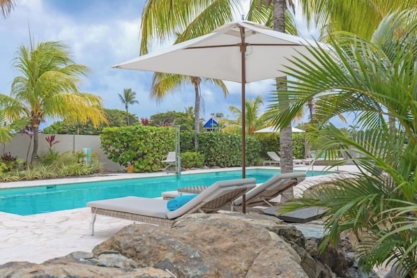 A pool area with lounge chairs and umbrellas, surrounded by palm trees and rocks. - Dolphin Suites (Willemstad)