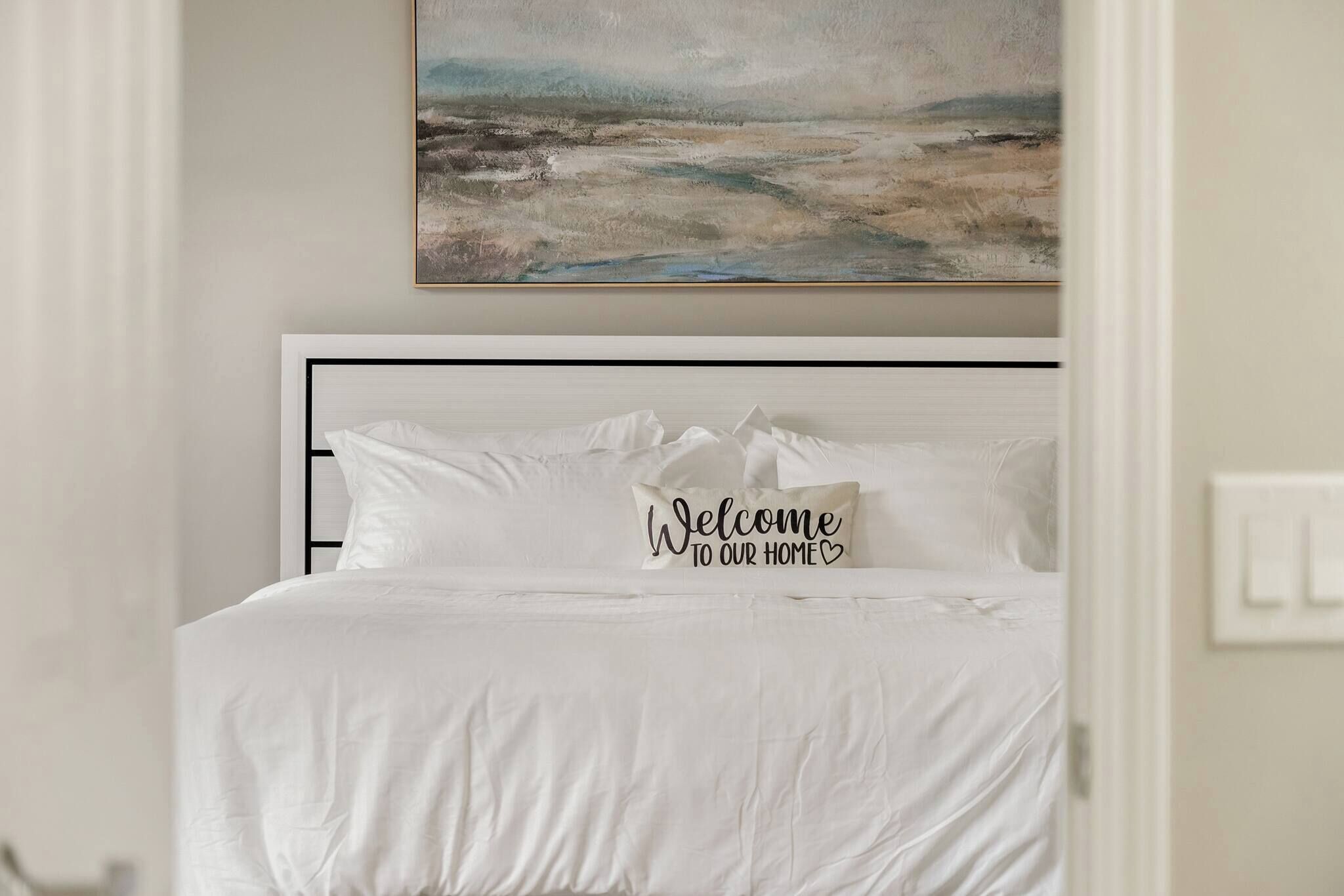 Un lit bien fait, avec une literie blanche et un coussin sur lequel est brodé le message : « Bienvenue chez nous ».