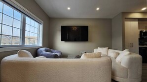 A modern living room with a large window, a flat-screen TV mounted on the wall, and a kitchen area with cabinets and appliances. - Harmony Haven in Drumheller (Drumheller)