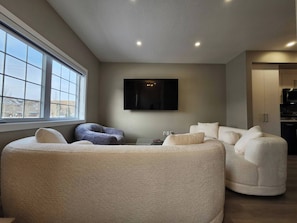 Een moderne woonkamer met een groot raam, een flatscreen televisie aan de muur en een keuken met kasten en apparatuur.