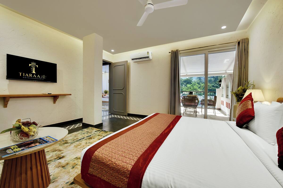 A hotel room with a bed, a desk with a magazine, a fruit bowl, a ceiling fan, and a balcony with a chair.