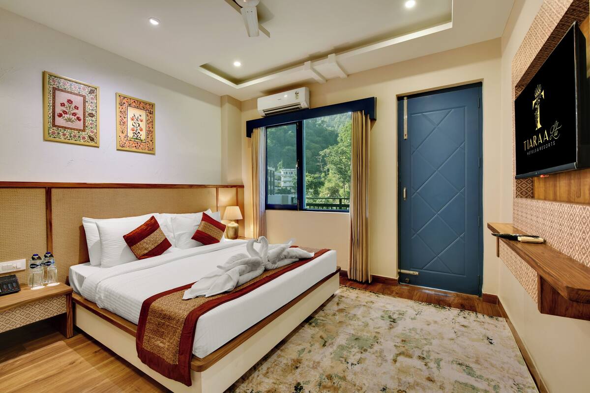 A hotel room with a large bed, a bedside table with a phone and glasses, a wall-mounted TV, and a window with curtains.