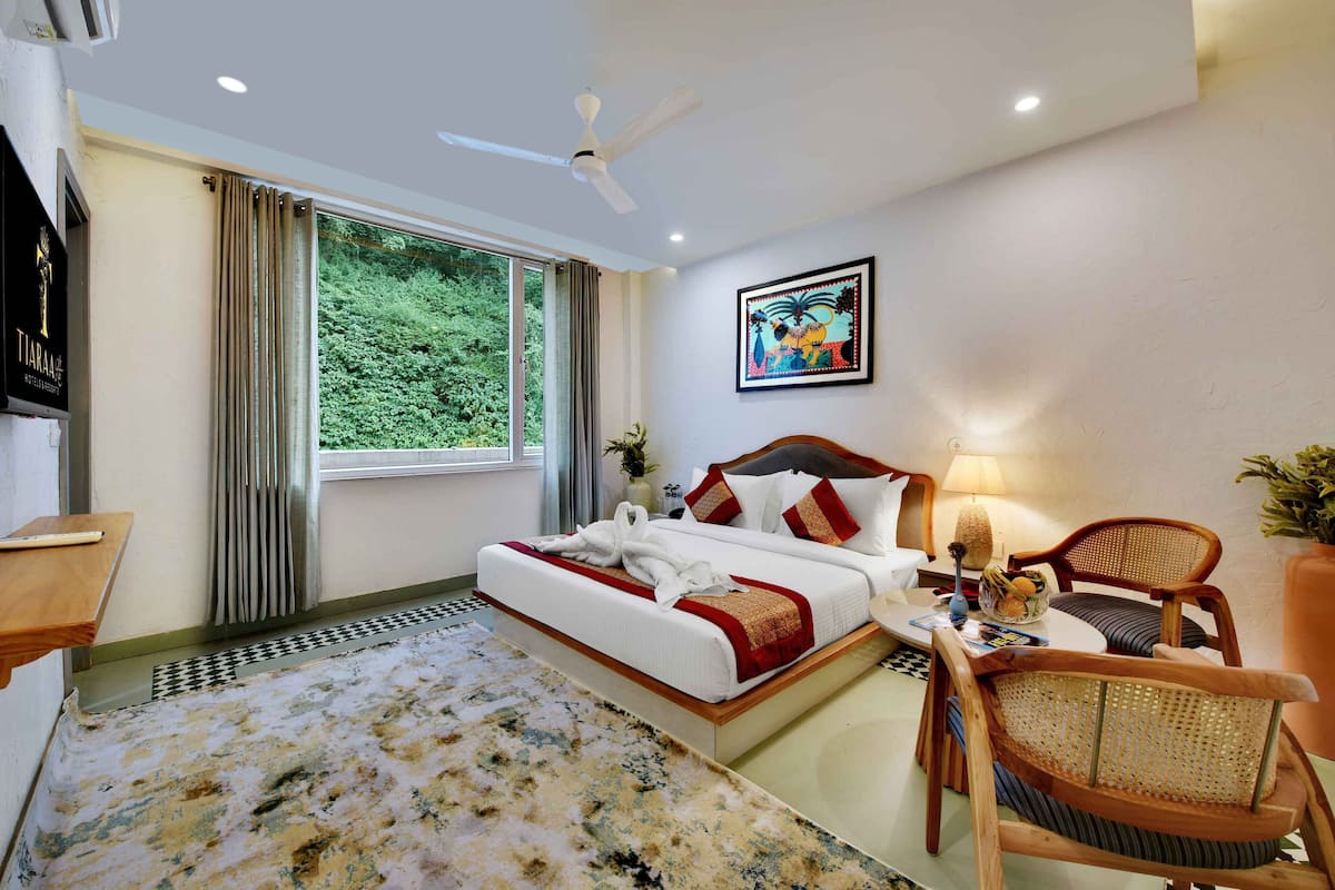 A hotel room with a bed, a desk, a chair, a ceiling fan, a window with curtains, a painting, and a view of greenery outside.