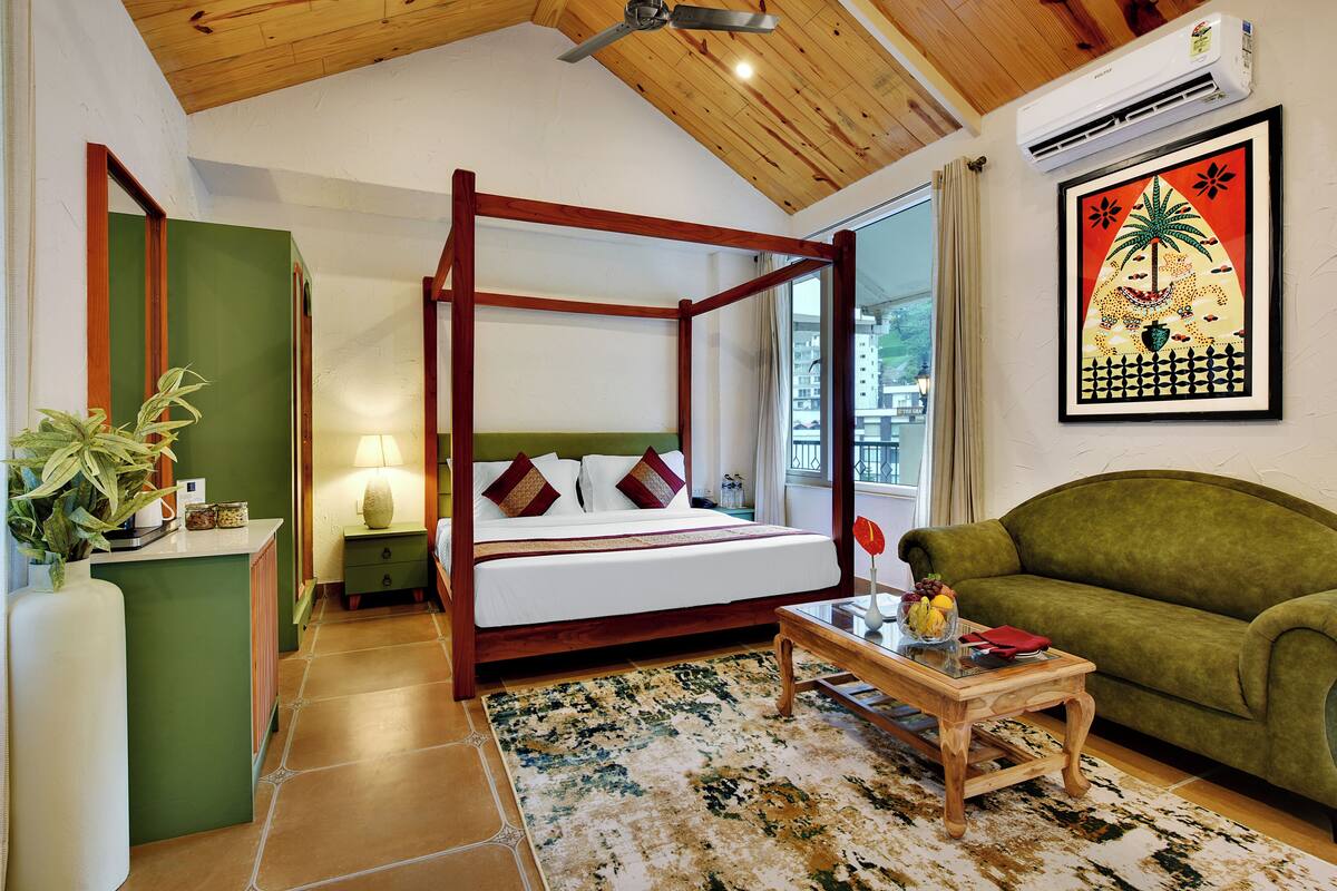 A four-poster bed with a wooden frame, a green sofa, a small table with a vase of flowers, and a framed picture on the wall.
