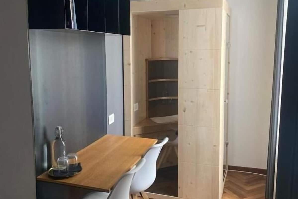 Een compacte kamer met een houten eettafel, een witte stoel, een inbouwkast en een kleine kitchenette.