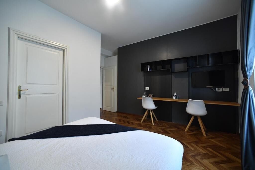 A bedroom with a bed, a desk, two chairs, a dark wall, and a door.