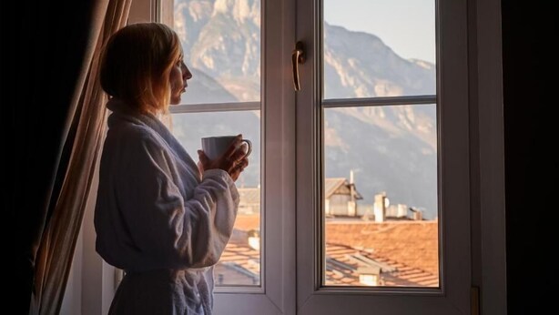 Eine Person in einem Bademantel am Fenster mit Blick auf Berge und Gebäude.