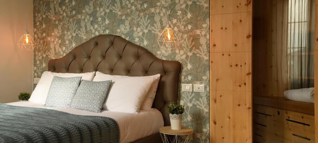 A bedroom with a tufted headboard, patterned wallpaper, a bedside plant, and a small round table.