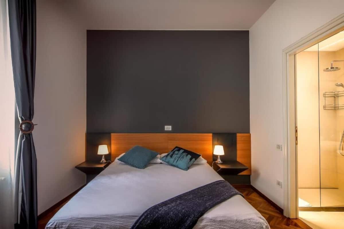 Een hotelkamer met een groot bed, twee nachtkastjes met lampen, een donker hoofdbord en een douchecabine met glazen wanden.
