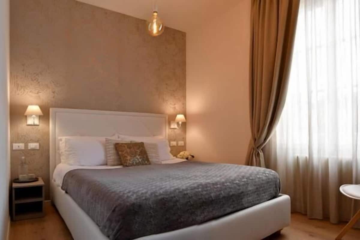 Een slaapkamer met een bed, nachtlampjes, een stoel en een raam met gordijnen.