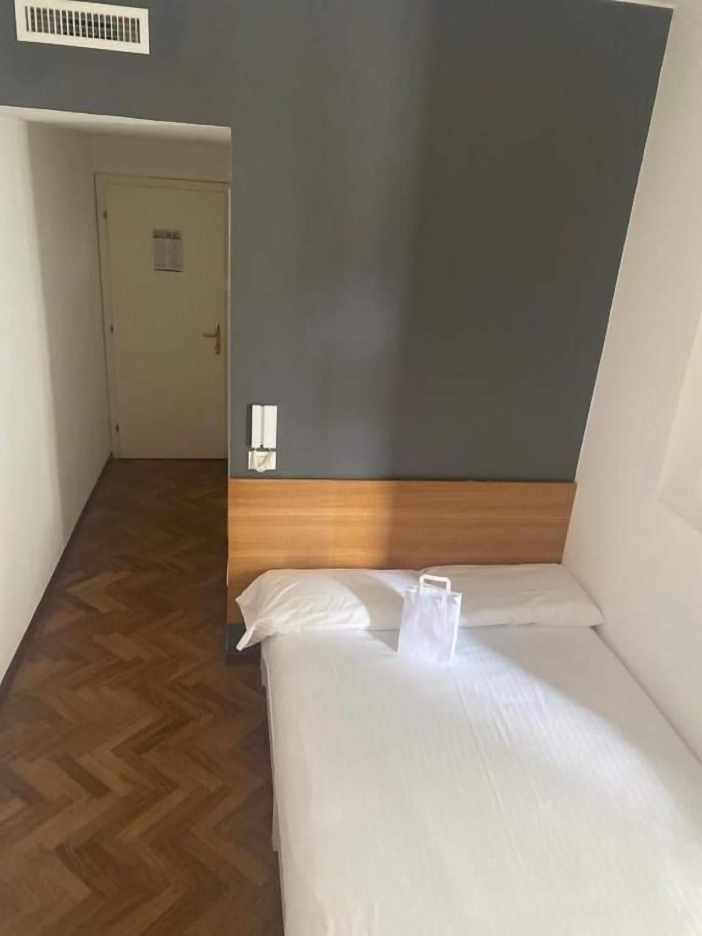 A small, single-bed room with a wooden headboard, a white bedspread, and a white pillow. A white bag is placed on the bed. The room has a wooden floor and a door leading to another room.